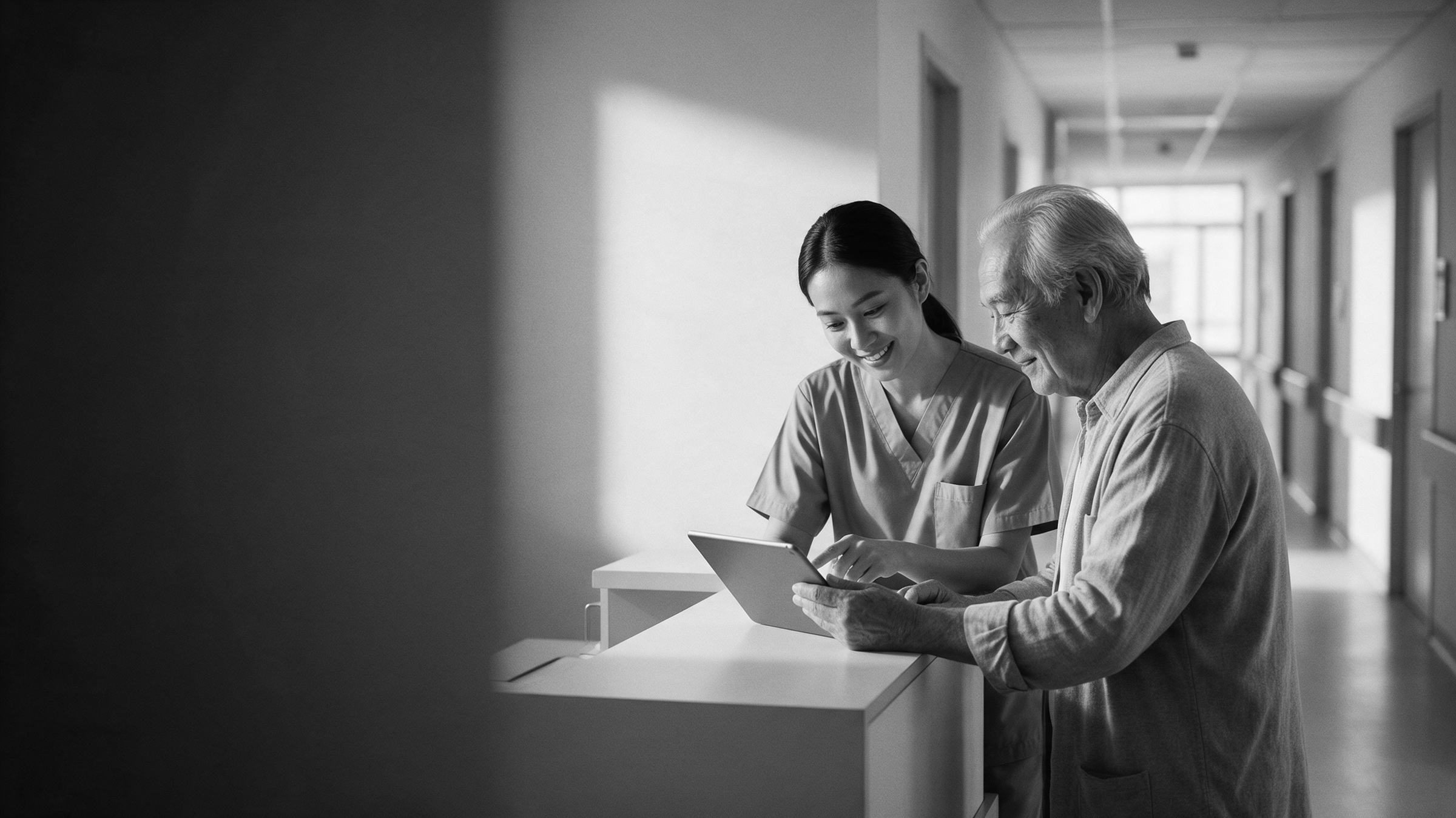 A nurse and an elderly patient reviewing a tablet in a bright hospital corridor